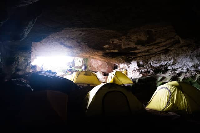 Vier gelbe Kuppelzelte in einer weitläufigen Felshöhle aufgebaut, während Wanderer am hell erleuchteten Höhleneingang stehen