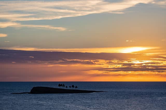 Silhueta de um pequeno ilhéu com palmeiras no arquipélago de Abrolhos contra um céu alaranjado e rosado ao pôr do sol