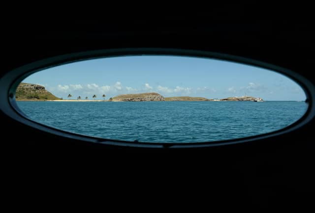 Vista através de uma vigia oval de embarcação para as ilhas Abrolhos emergindo das águas azuis do Atlântico, com coqueiros, morros rochosos e um farol ao fundo em um passeio pelos Abrolhos