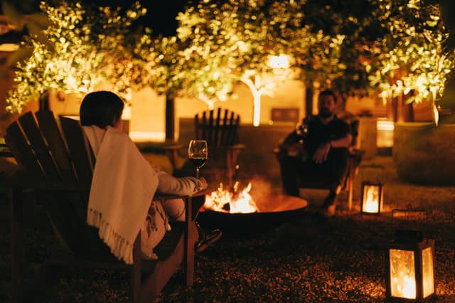 Twee gasten genieten van een luxe rondreis door Brazilië bij een gloeiende vuurschaal in de nacht, ingepakt in dekens met rode wijn en lantaarns onder verlichte bomen