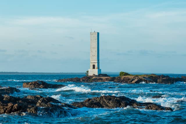 Ein hoher Betonleuchtturm steht auf einem felsigen Inselchen, umgeben von tosenden blauen Atlantikwellen nahe Itacaré, Bahia, Brasilien