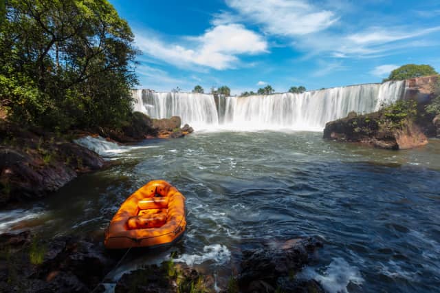 Ein orangefarbenes Schlauchboot am felsigen Flussufer vor einem breiten Wasserfall im Cerrado-Wald auf einer Jalapão-Tour in Brasilien