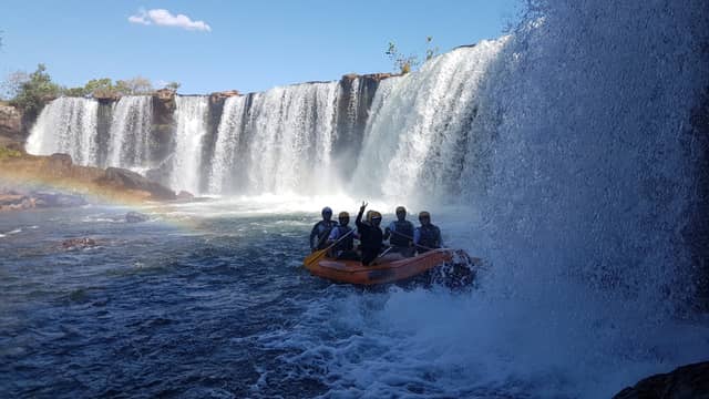 Grupo de rafters capacetados em bote inflável laranja remando pela névoa intensa na base de uma cachoeira larga no Jalapão, com um arco-íris visível na neblina