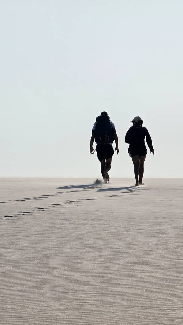 Zwei Trekker mit Rucksäcken überqueren eine weite, vom Wind gekräuselte Sandfläche und hinterlassen eine Spur von Fußabdrücken