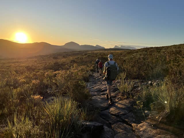 A group of hikers on the vale do pati trek walking single-file along a rocky trail through cerrado scrubland toward a vivid golden sunset, with a panorama of Chapada Diamantina mountains stretching to the horizon