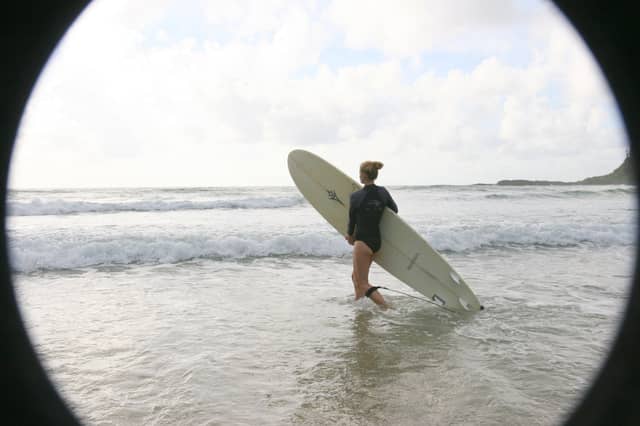 A woman in a black wetsuit top carries a longboard into the surf during a surfing in Brazil session, seen through a circular vignette frame with rolling waves and a headland in the background.