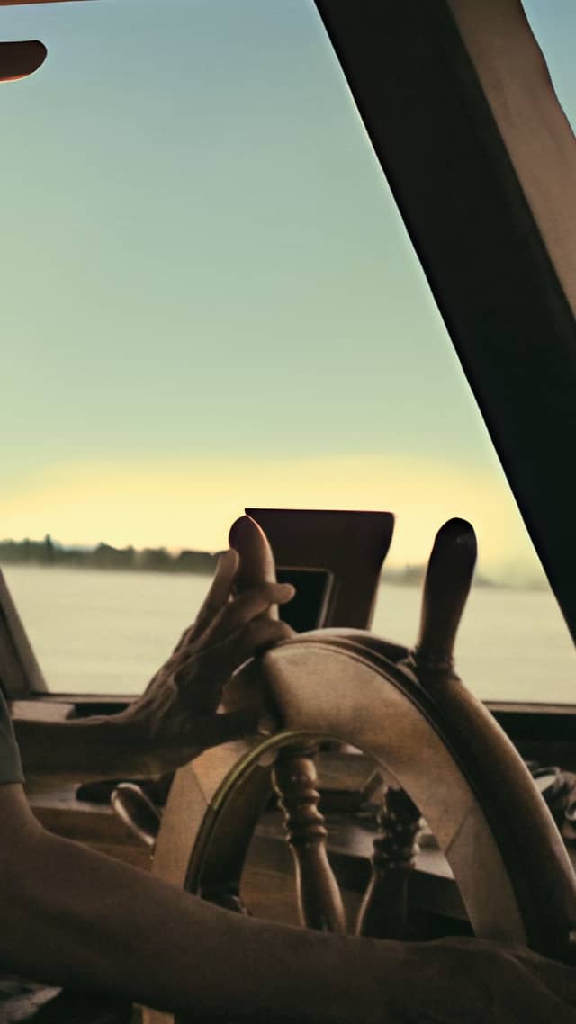 A hand rests on a wooden boat helm during a boat tour in Brazil, with a wide calm river and pale dawn sky visible through the windscreen