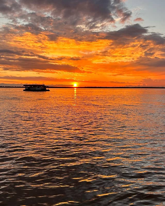 A houseboat silhouetted against a blazing orange and red sunset reflected across a wide Amazonian river, the kind of scene encountered on an Amazon trek and survival tour departing from Manaus