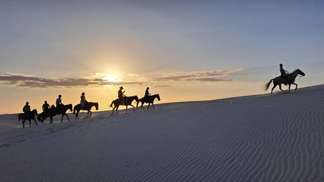 Silhouettes of seven riders on horseback crossing white sand dunes during a golden sunset on a brazil group tours adventure