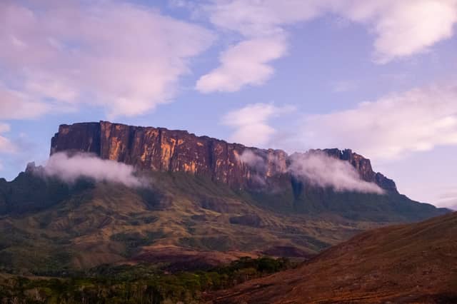 De steile zandstenen kliffen van Mount Roraima kleuren amberkleurig in het avondlicht, omgeven door lage wolken boven het Gran Sabana-hoogland