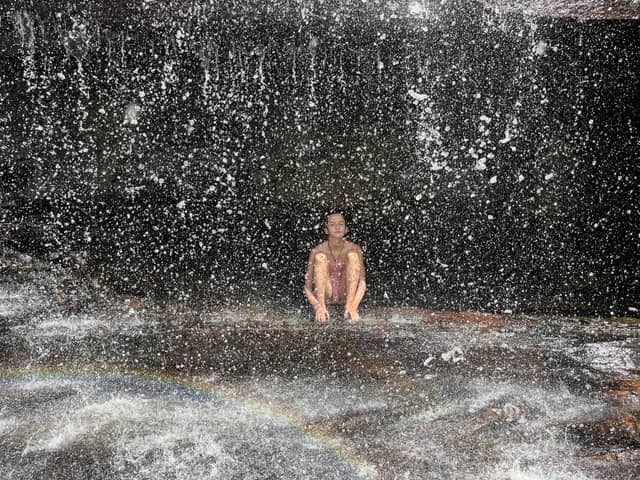 A traveller sits eyes closed on a wet rock ledge beneath a cascading waterfall on the Pati Valley hike, with droplets filling the air and a small rainbow forming at the base
