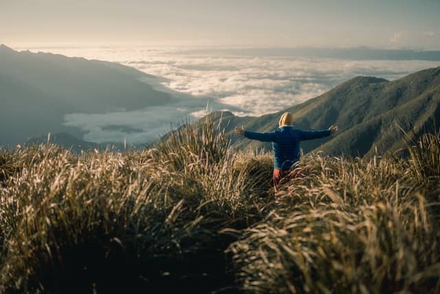 A hiker in a blue jacket stands arms wide open on a Serra Fina Trekking ridge, surrounded by golden grass with a sea of clouds filling the Mantiqueira valleys below