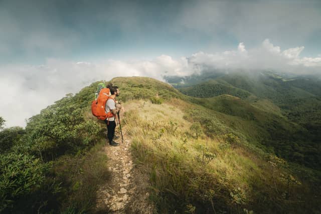 A hiker with an orange backpack and trekking poles pauses on a narrow exposed ridge during a Serra Fina Trekking traverse, with rolling cloud-covered peaks of the Serra da Mantiqueira stretching to the horizon