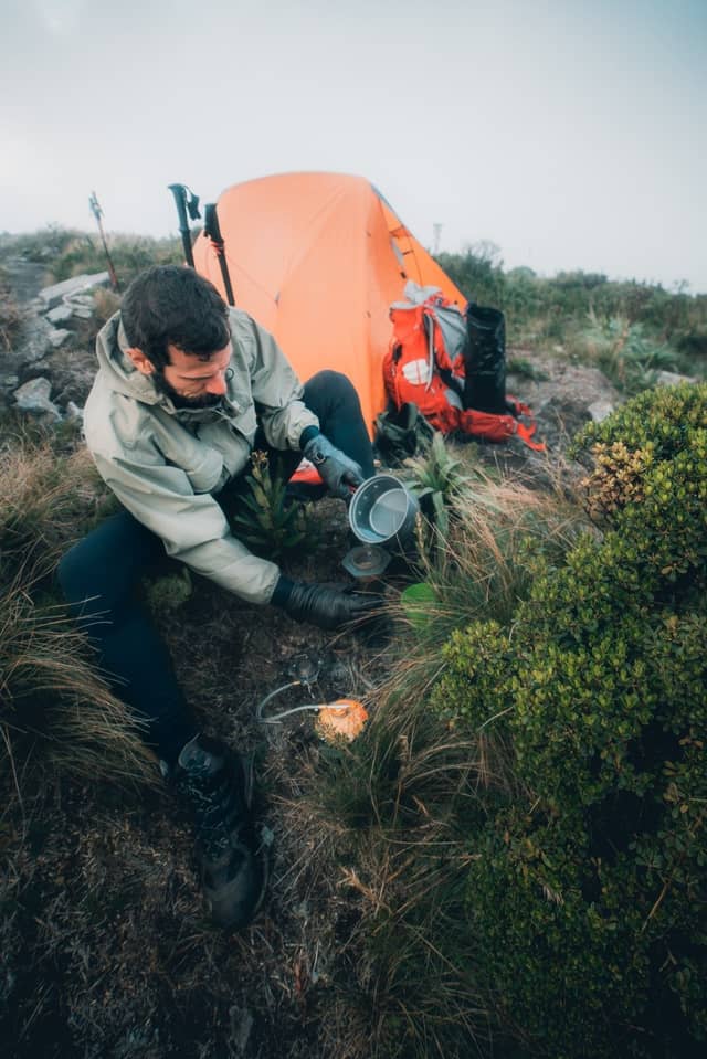 A bearded hiker in a rain jacket pours water into a pot on a compact camp stove beside an orange tent and red backpack during a Serra Fina Trekking expedition in the Serra da Mantiqueira highlands