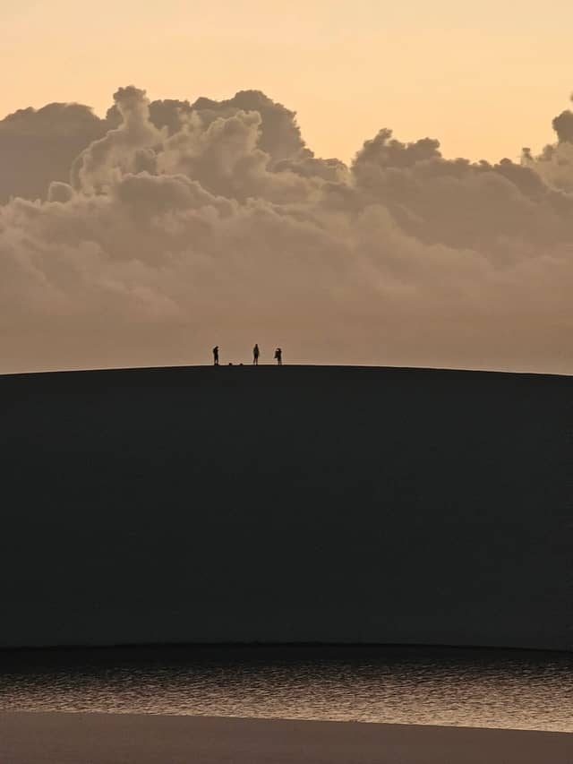 Three tiny silhouettes stand on the curved ridge of a vast sand dune at golden-hour dusk, a dramatic cumulus cloudbank rising behind them — a defining moment in trekking in Brazil's Lençóis Maranhenses.