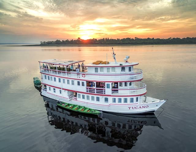 The M/Y Tucano amazon river cruise vessel moored on glassy dark water at sunset, with guests on the upper observation deck and a green skiff alongside, forested horizon silhouetted against an orange sky