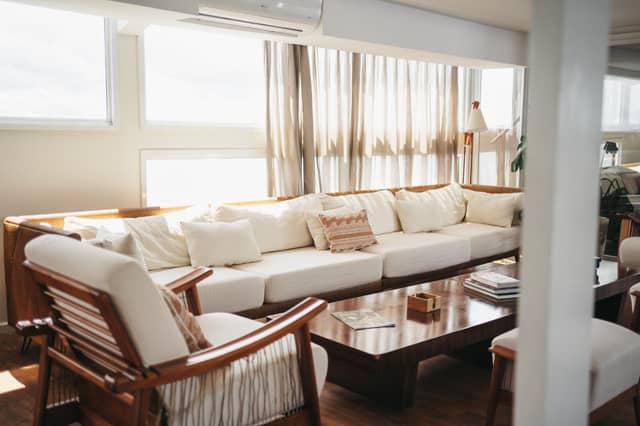 Sunlit social lounge on the untamed amazon cruise featuring a wide cream sofa with rattan frame, wooden armchairs, and a polished coffee table beneath sheer linen curtains