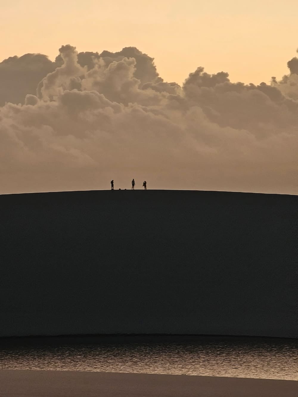 Silhouettes of three people walking on top of a sand dune in Lencois Maranhenses during sunrise, with clouds in the background.