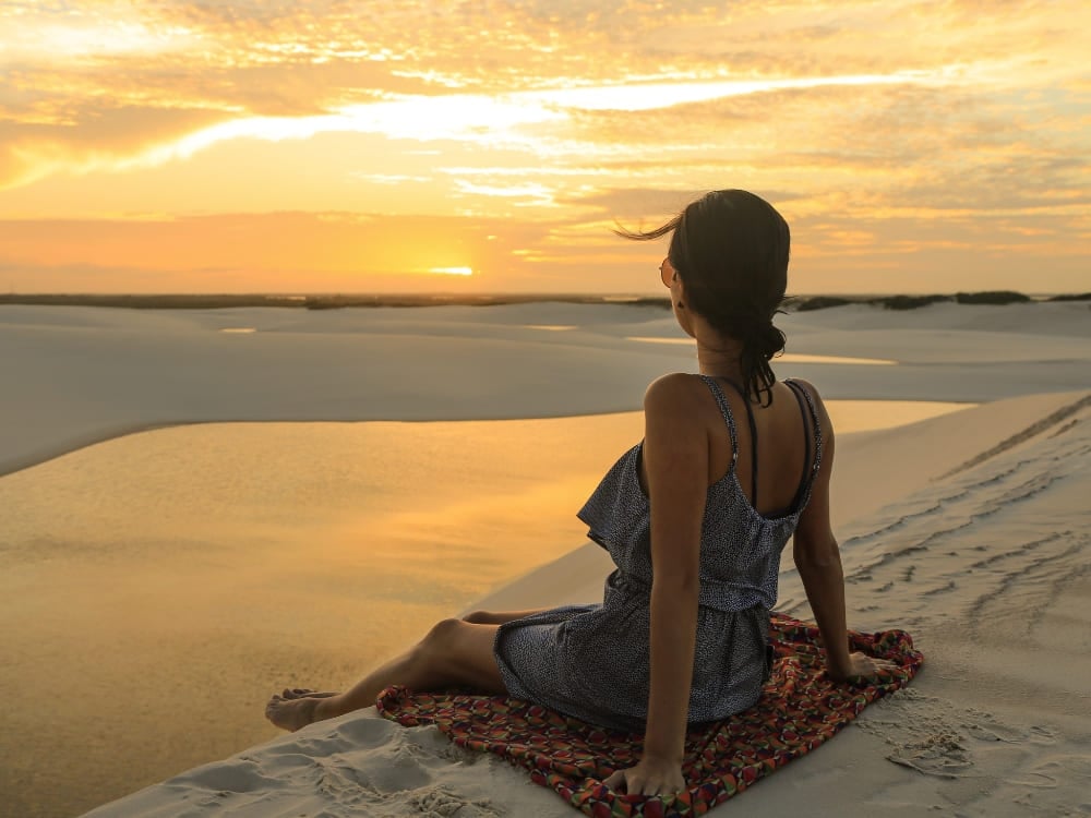 Mujer sentada sobre un pareo en la arena blanca, admirando el atardecer dorado que se refleja en las aguas de las lagunas de Lençóis Maranhenses.