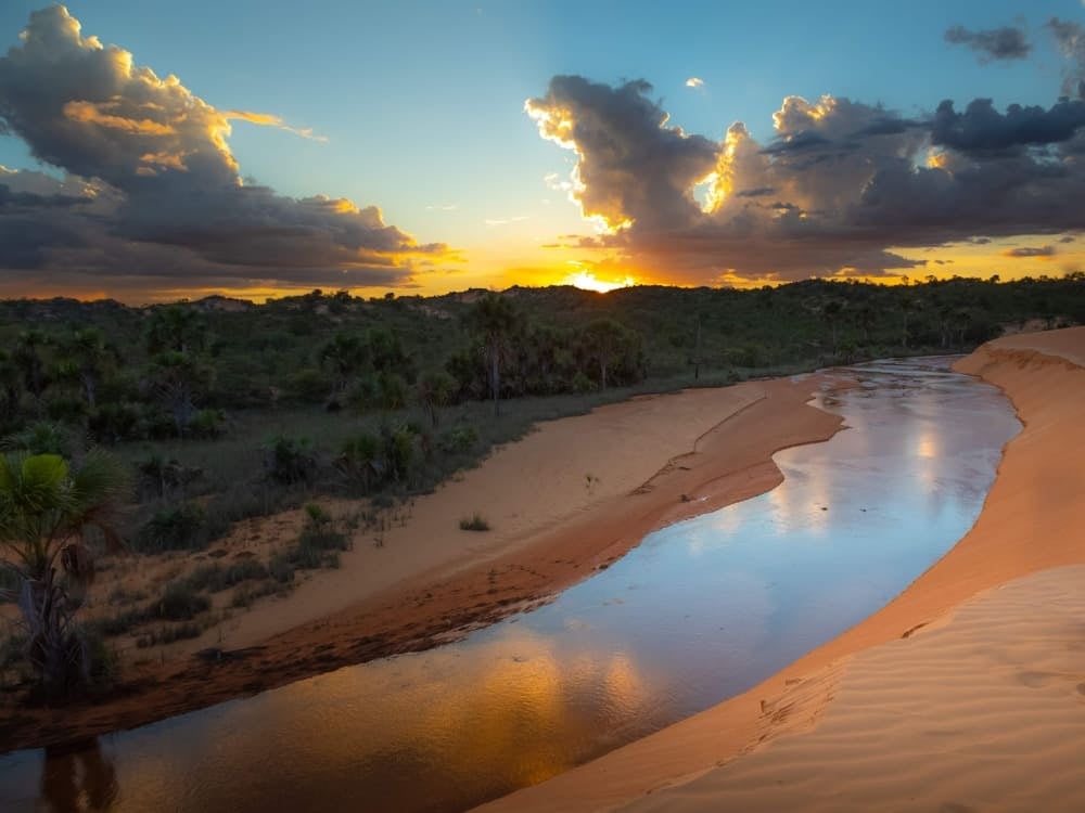 Un fiume calmo scorre fra ampie distese di sabbia e falesie frastagliate al tramonto, e riflette la regione