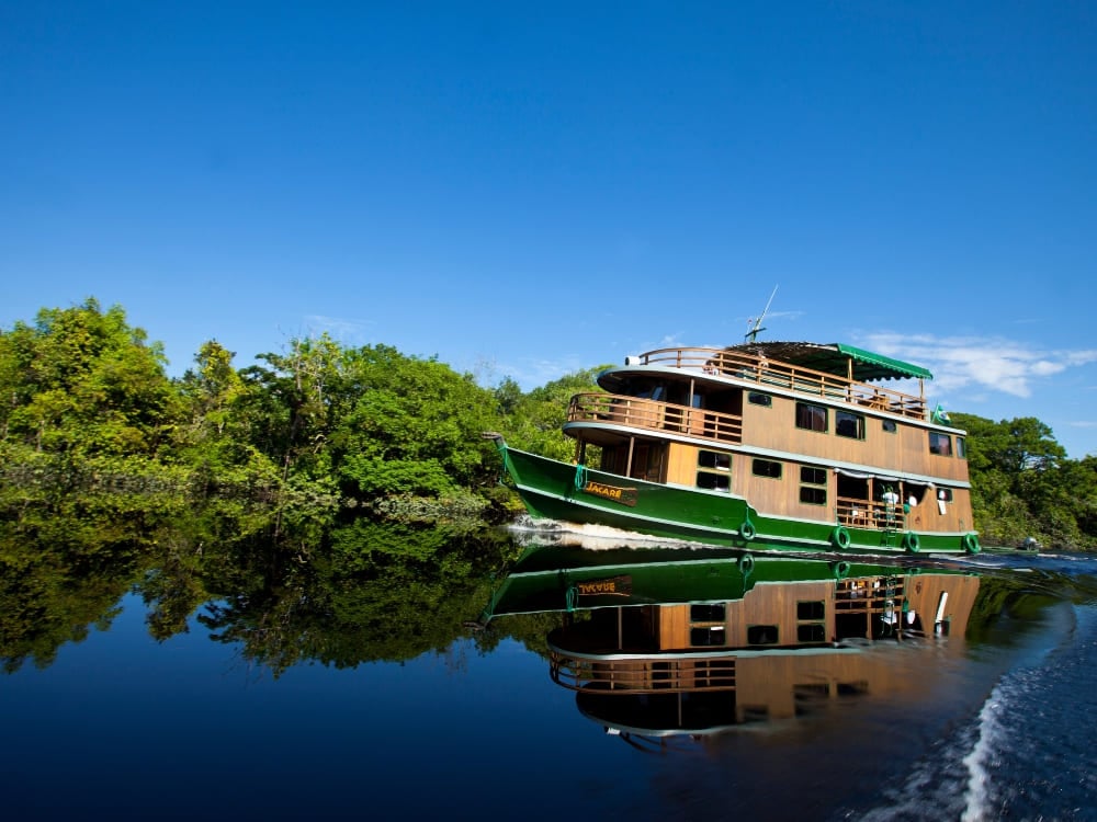 Una rustica barca fluviale di legno a tre ponti naviga tranquilla su un fiume amazzonico scuro sotto un cielo azzurro.