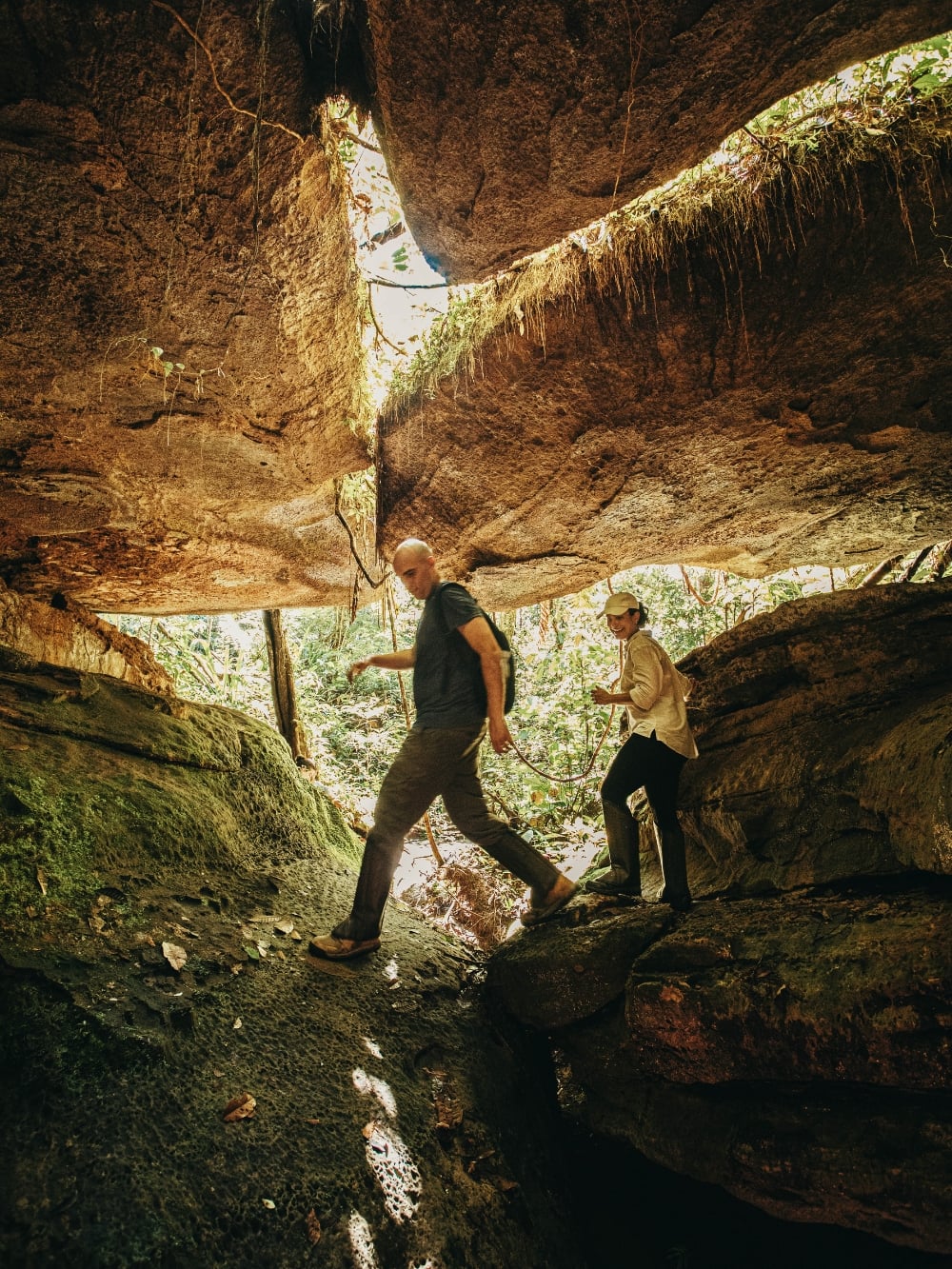 Two travelers hiking under a massive rock formation in the Presidente Figueiredo forest.