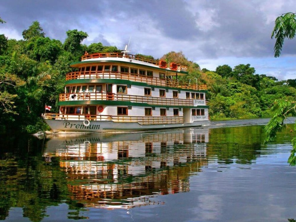 La Amazon Clipper Premium naviga lungo il Rio delle Amazzoni e si riflette nell'acqua calma, sullo sfondo della foresta pluviale fitta e verde.