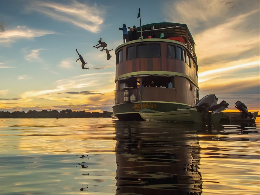 Viaggiatori si tuffano dal ponte superiore di una barca da crociera nel Rio delle Amazzoni al tramonto, durante un viaggio di ecoturismo avventuroso