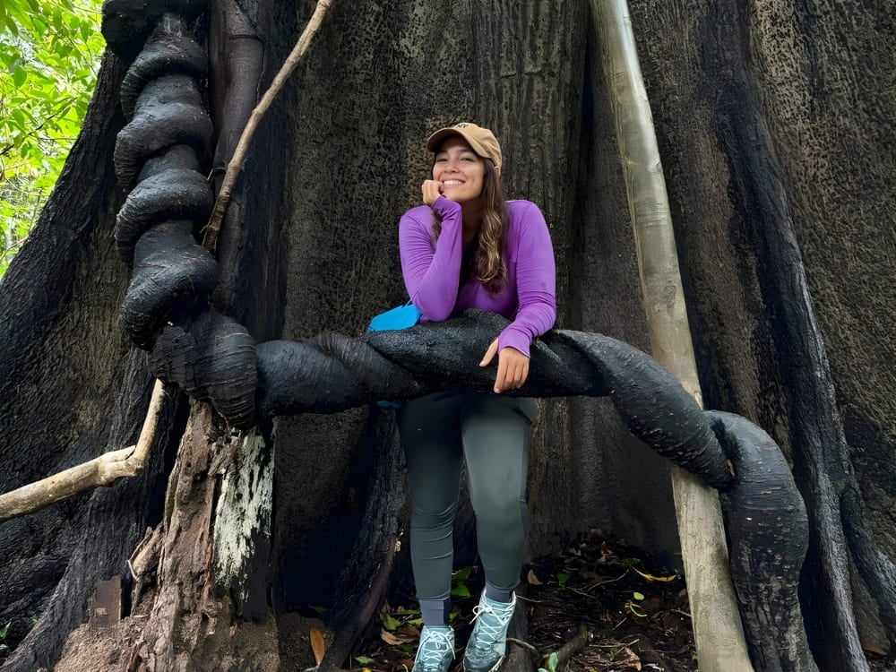 Une femme souriante portant un chemisier violet, appuyée sur les racines épaisses et tordues d'un arbre géant au milieu d'une forêt d'un vert luxuriant.