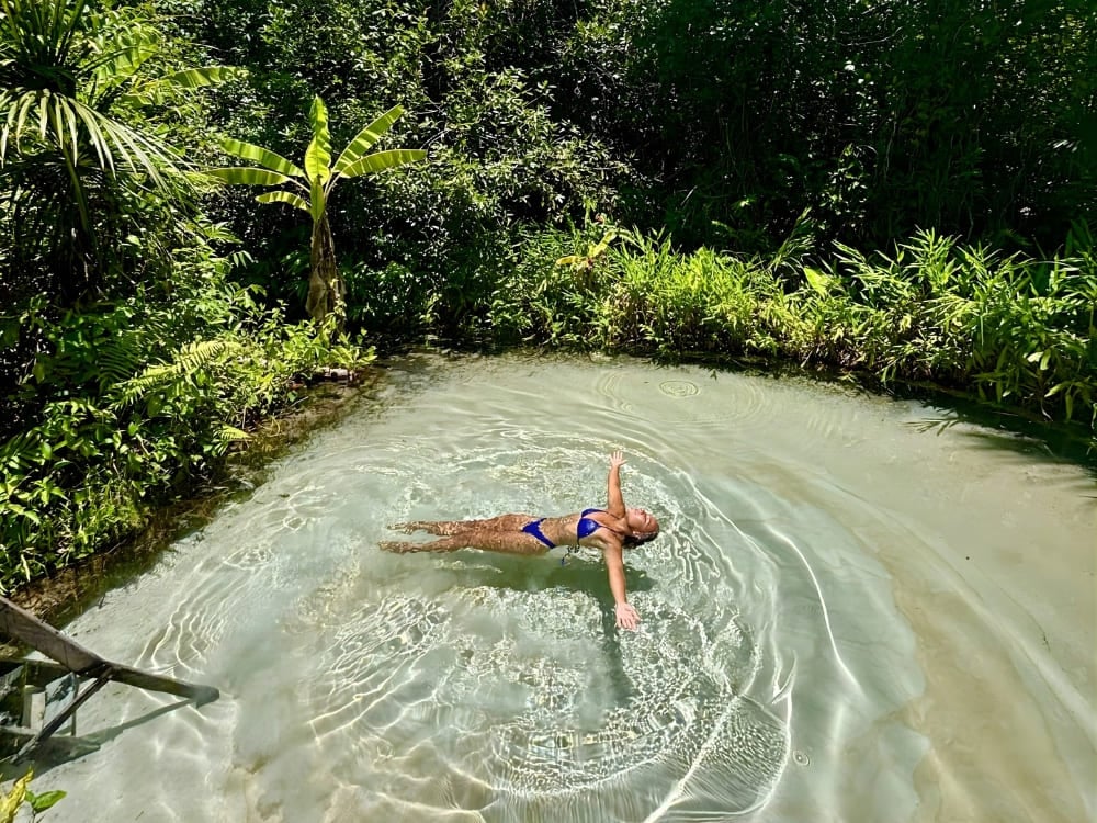 Une femme flottant sur le dos, les bras ouverts, dans des eaux cristallines d'un bleu clair, entourée d'arbres tropicaux et d'une végétation verdoyante et dense.