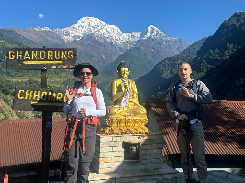 Una donna e un ragazzo con zaini da trekking sorridono accanto a una statua dorata del Buddha, con le maestose montagne innevate dell'Himalaya sullo sfondo.