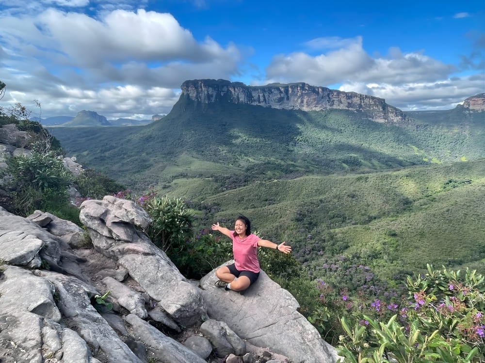 Une femme souriante assise en tailleur et les bras ouverts sur un rocher, admirant une vallée verdoyante avec une imposante montagne au sommet plat en arrière-plan.