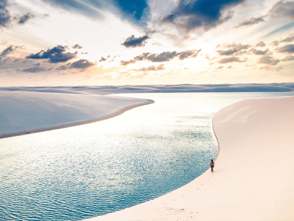 A single person walks along the edge of a vibrant blue lagoon winding through vast white sand dunes under a dramatic cloudy sky. A single person walks along the edge of a vibrant blue lagoon winding through vast white sand dunes under a dramatic cloudy sky.