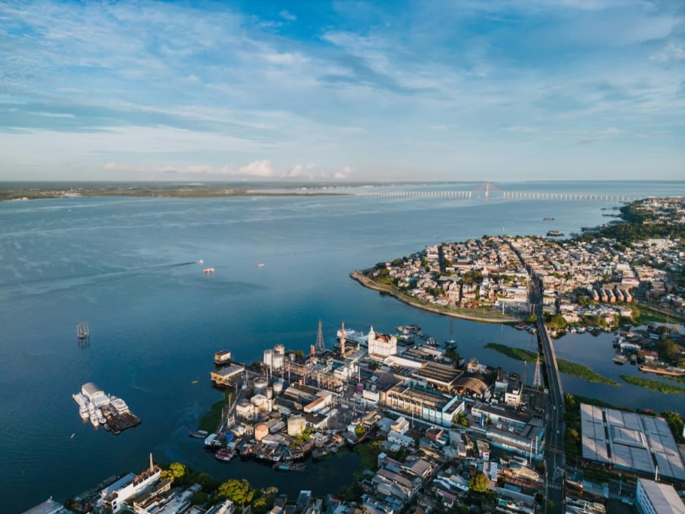 Aerial view of Manaus, Amazonas, showing the city's port, residential neighborhoods, and the expansive river with a long bridge in the distance.