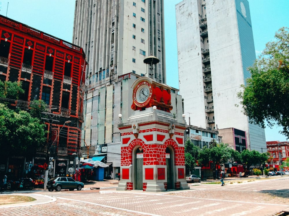 Historic red and stone clock tower standing in a city square against a backdrop of modern tall buildings in Manaus.