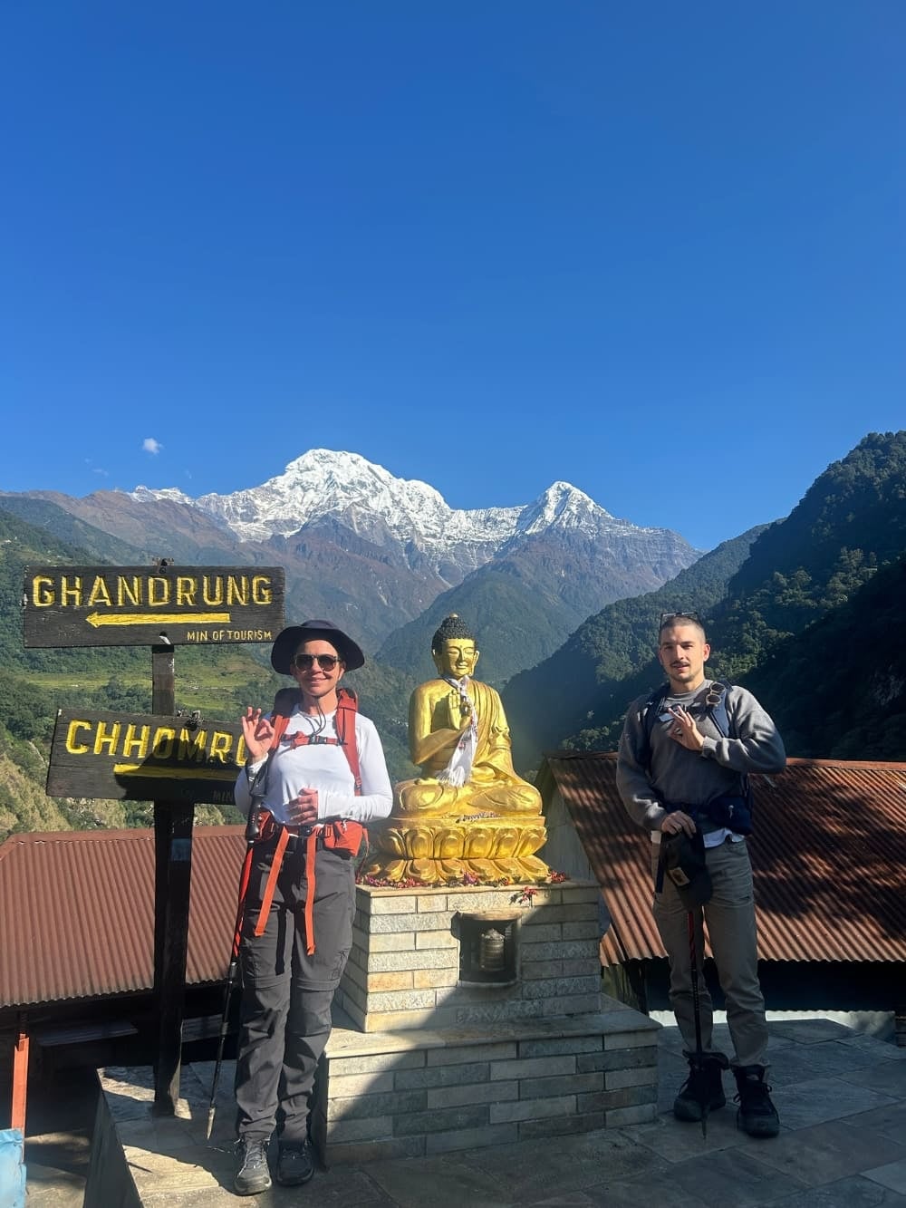 Mila und Lorenzo neben einer Buddha-Statue und Wegweisern während ihres nachhaltigen Trekkings in Nepal.