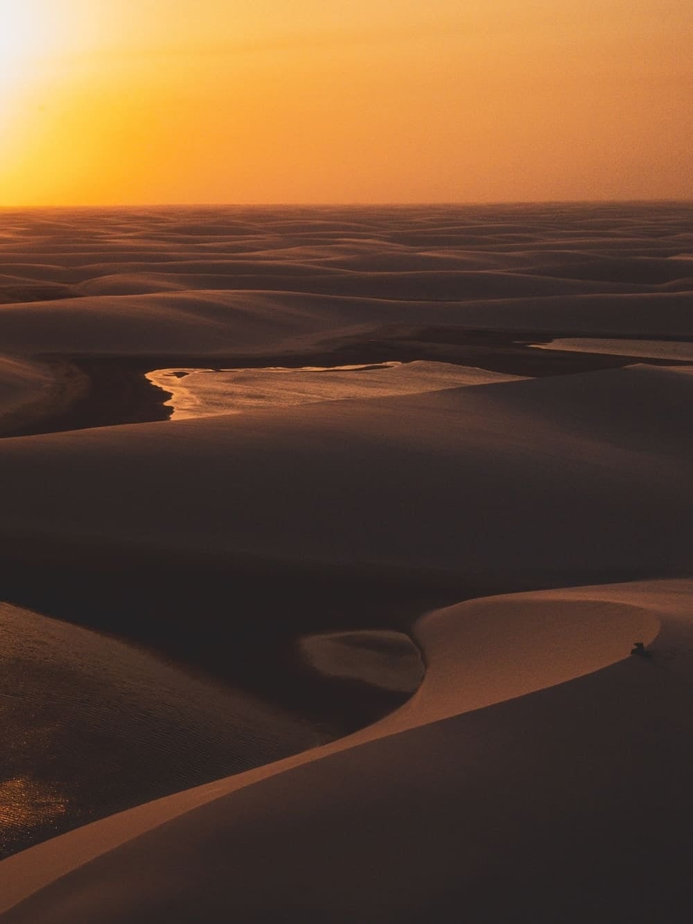 Vasta distesa di dune lisce illuminate da un tramonto dorato nel Parco Nazionale dei Lençóis Maranhenses, in Brasile. Una pozza d'acqua bassa riposa nella valle tra le dune e riflette il cielo arancione.