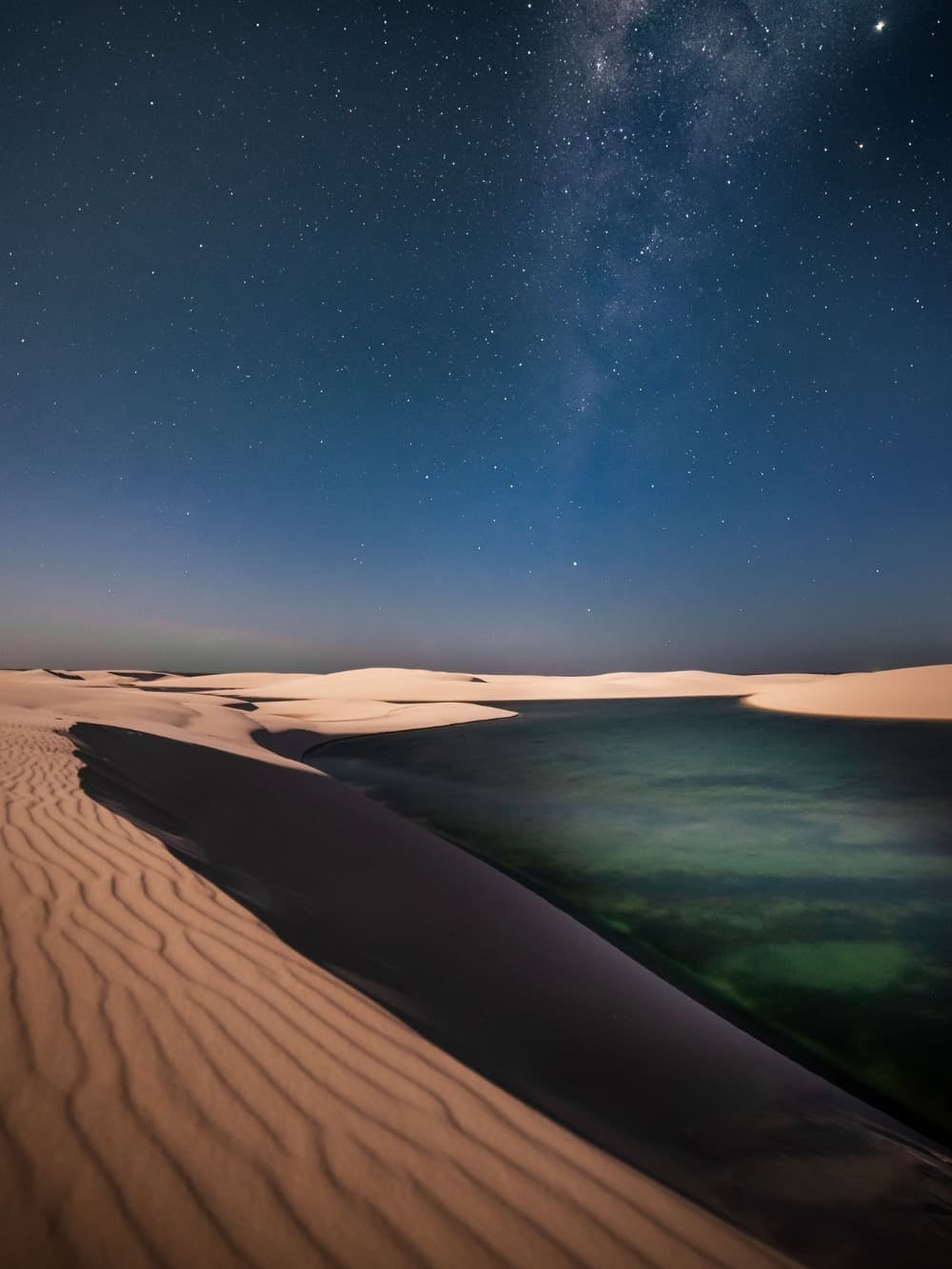 Paesaggio notturno di dune nei Lençóis Maranhenses, in Brasile, sotto un cielo scuro pieno di stelle, con la Via Lattea ben visibile. In primo piano una laguna scura con riflessi verdi e blu ai piedi di una duna increspata.