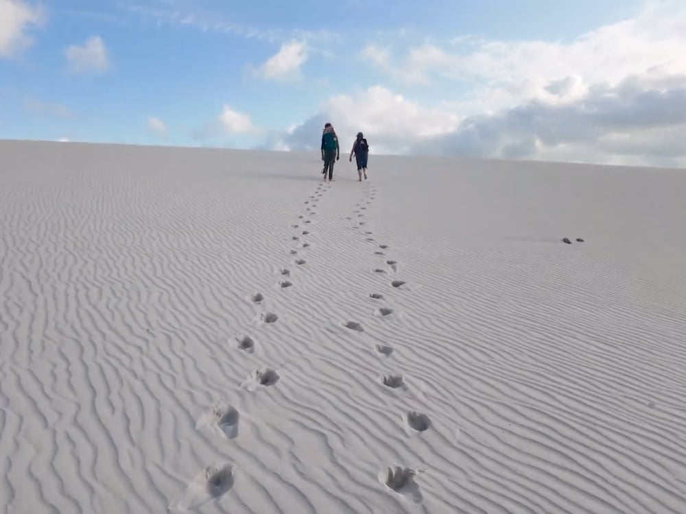 Two trekkers crossing a rippled white dune in Lençóis Maranhenses, footprints trailing behind them