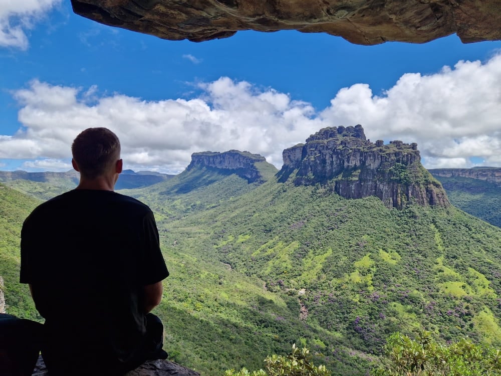 Un uomo seduto sotto uno strapiombo di roccia osserva un'ampia valle verde e le montagne dalla cima piatta della Chapada Diamantina. Un uomo seduto sotto uno strapiombo di roccia osserva un'ampia valle verde e le montagne dalla cima piatta della Chapada Diamantina.