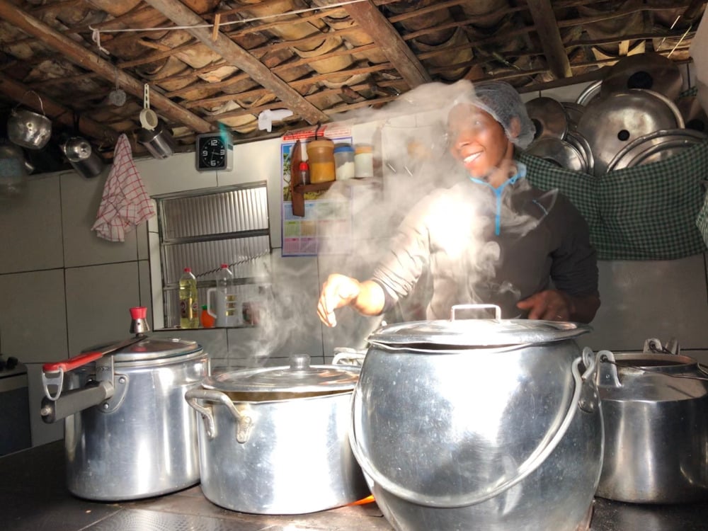 Eine einheimische Frau lächelt, während sie mit großen Aluminiumtöpfen auf einem Herd eine traditionelle hausgemachte Mahlzeit kocht. Eine einheimische Frau lächelt, während sie mit großen Aluminiumtöpfen auf einem Herd eine traditionelle hausgemachte Mahlzeit kocht.