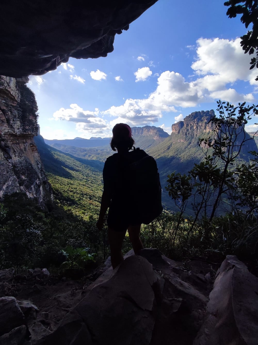 Silhouette eines Wanderers mit Rucksack, der an einem dunklen Höhleneingang steht und über das sonnenbeschienene Pati-Tal blickt. Silhouette eines Wanderers mit Rucksack, der an einem dunklen Höhleneingang steht und über das sonnenbeschienene Pati-Tal blickt.