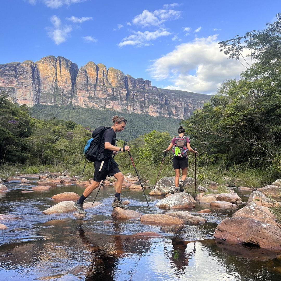 Zwei Wanderer nutzen Trekkingstöcke, um einen flachen, felsigen Fluss zu überqueren, während sich im Hintergrund ein großer Berggipfel erhebt. Zwei Wanderer nutzen Trekkingstöcke, um einen flachen, felsigen Fluss zu überqueren, während sich im Hintergrund ein großer Berggipfel erhebt.