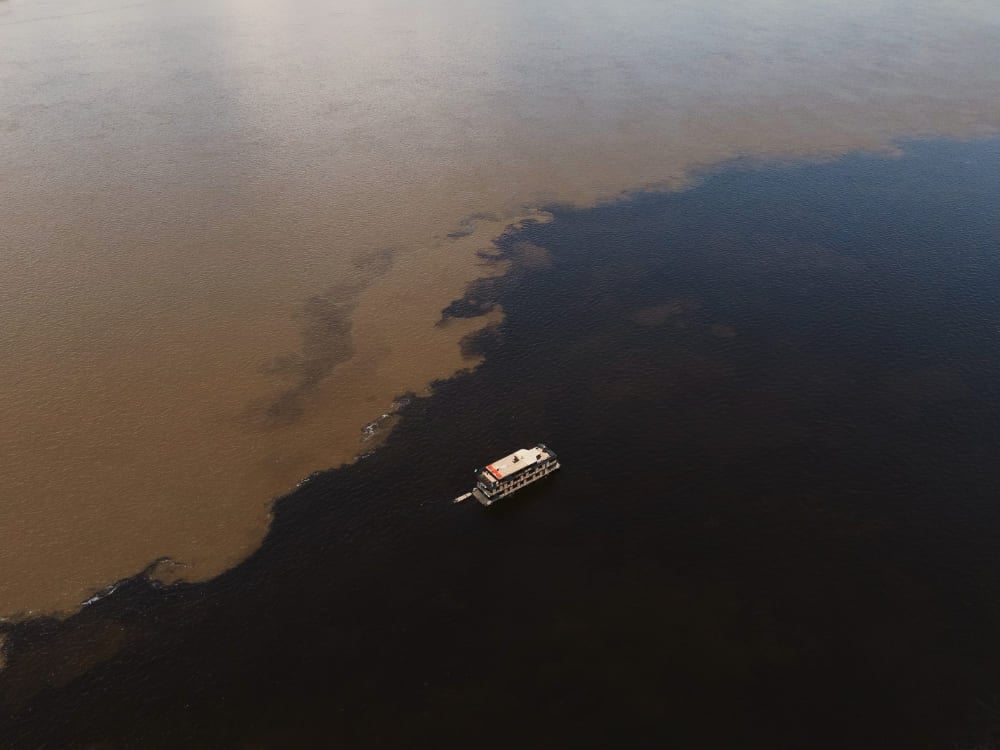 Veduta aerea di un battello che naviga sull'Incontro delle Acque, in Amazzonia, con i due fiumi ben distinti, uno scuro e uno color sabbia, che scorrono affiancati. Veduta aerea di un battello che naviga sull'Incontro delle Acque, in Amazzonia, con i due fiumi ben distinti, uno scuro e uno color sabbia, che scorrono affiancati.