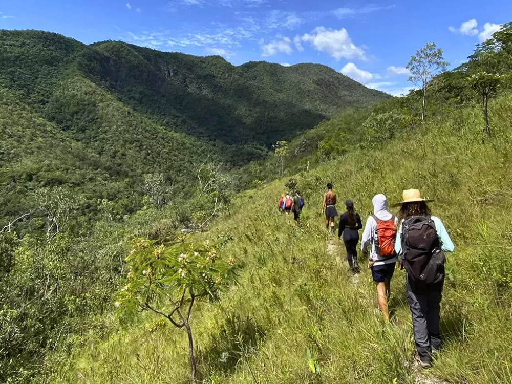 Eine Gruppe von Reisenden wandert im Gänsemarsch entlang eines schmalen, grasbewachsenen Pfades, der von den üppig grünen Tälern des Nationalparks Chapada dos Veadeiros umgeben ist. Eine Gruppe von Reisenden wandert im Gänsemarsch entlang eines schmalen, grasbewachsenen Pfades, der von den üppig grünen Tälern des Nationalparks Chapada dos Veadeiros umgeben ist.