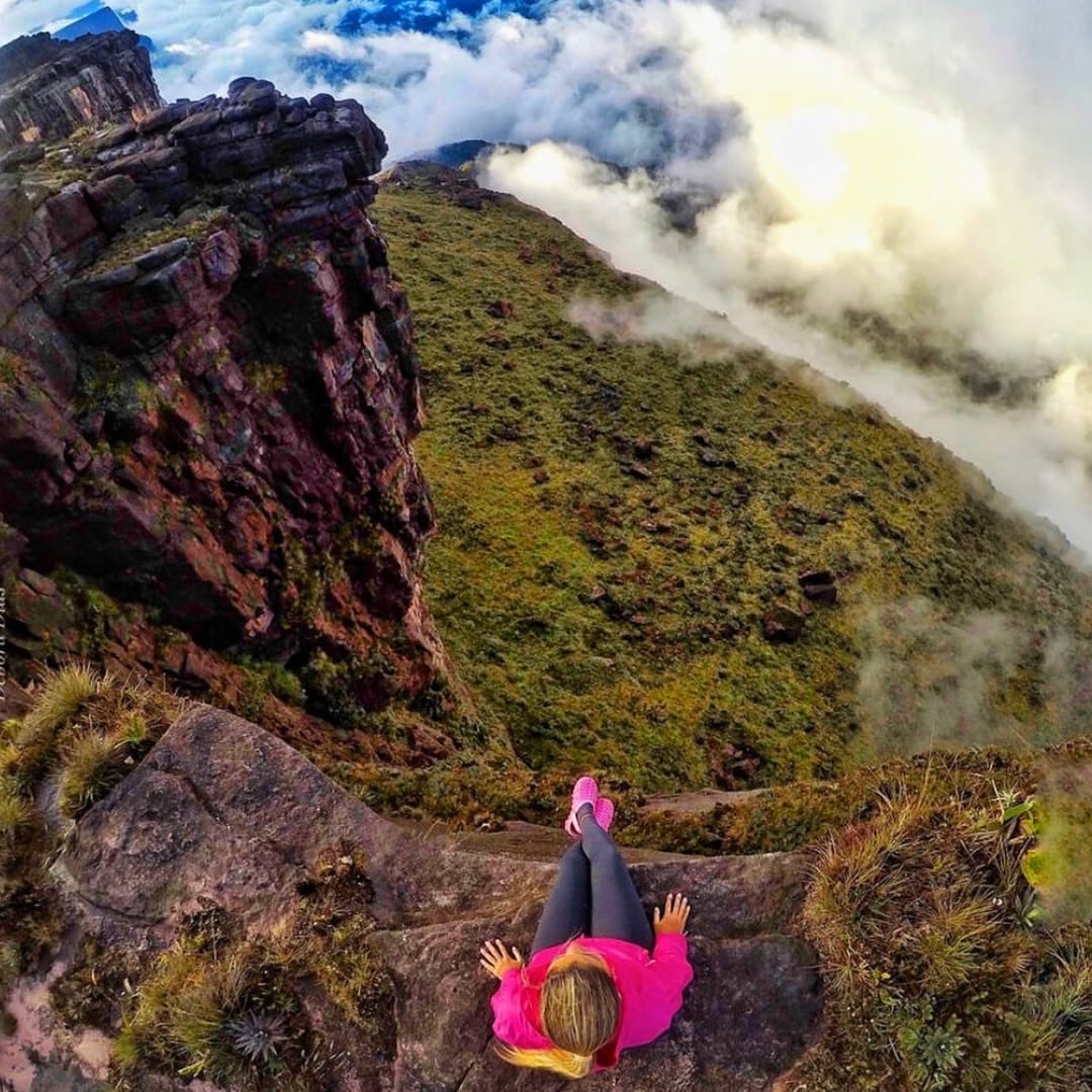 Ein Wanderer sitzt am Rand einer steilen, felsigen Sandsteinklippe am Monte Roraima und blickt auf ein weites, mit dichten weißen Wolken gefülltes Tal hinab. Ein Wanderer sitzt am Rand einer steilen, felsigen Sandsteinklippe am Monte Roraima und blickt auf ein weites, mit dichten weißen Wolken gefülltes Tal hinab.
