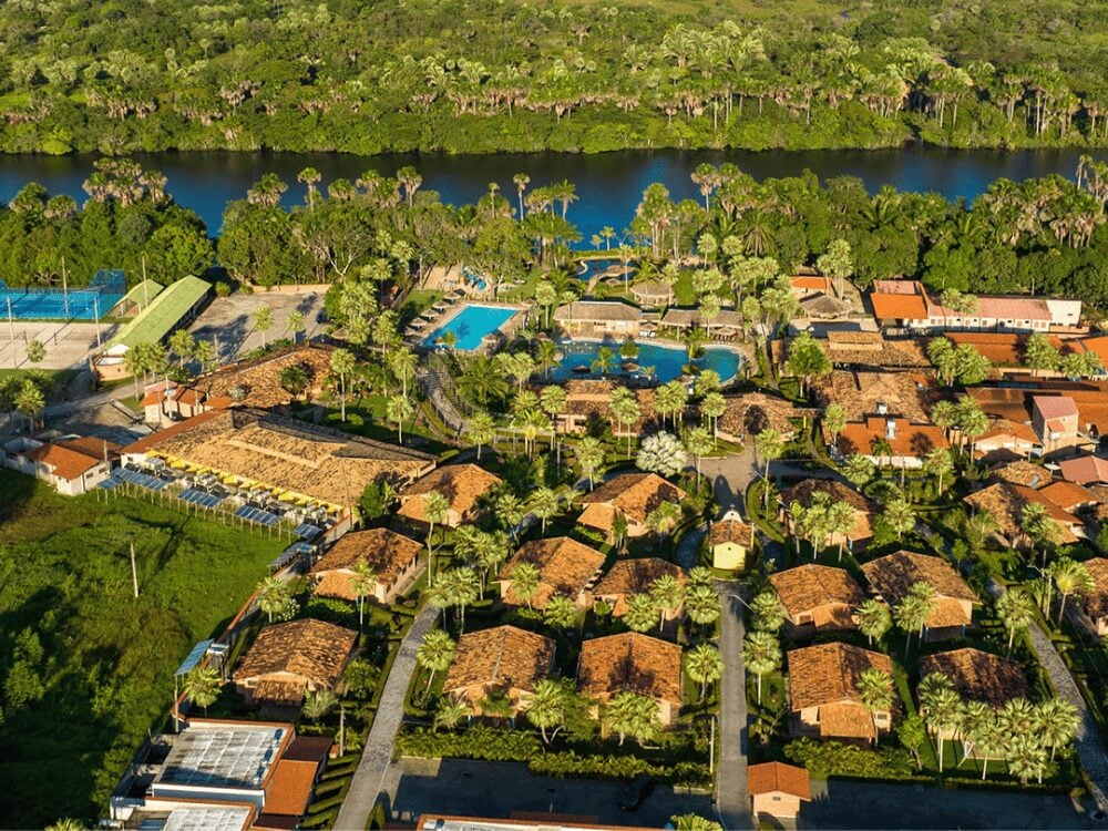 Aerial view of a lush resort complex featuring red-roofed buildings and blue swimming pools nestled beside a wide river. Aerial view of a lush resort complex featuring red-roofed buildings and blue swimming pools nestled beside a wide river.