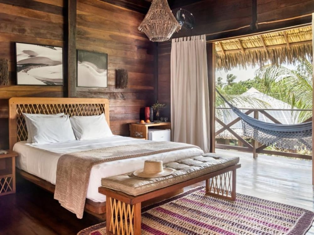 Camera accogliente rivestita di pannelli di legno scuro alla Charme Atins, con letto matrimoniale e porte aperte su un balcone con amaca. Camera accogliente rivestita di pannelli di legno scuro alla Charme Atins, con letto matrimoniale e porte aperte su un balcone con amaca.