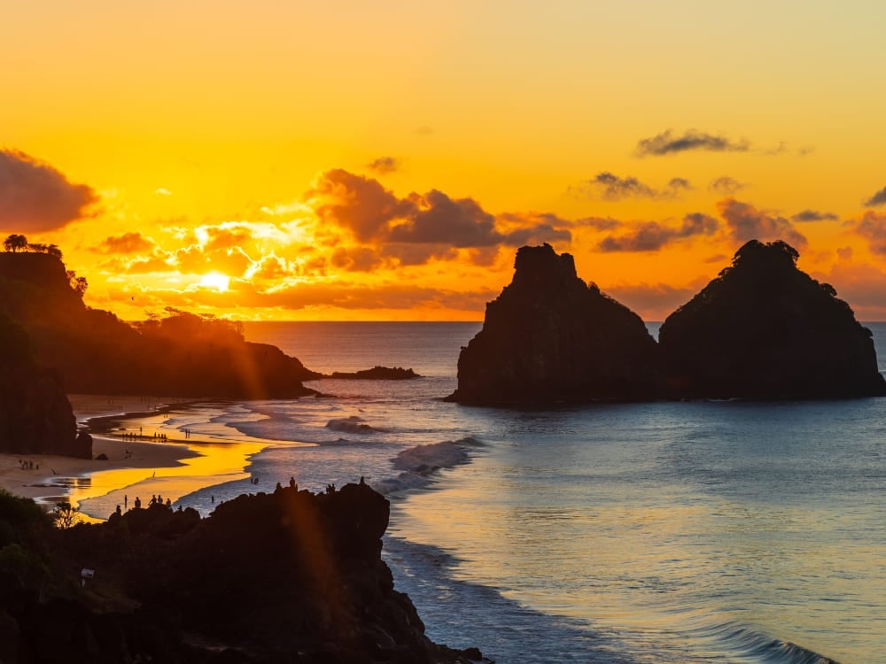 Un tramonto dorato illumina le onde e le formazioni rocciose del Morro dos Dois Irmãos mentre i surfisti pagaiano in acqua. Un tramonto dorato illumina le onde e le formazioni rocciose del Morro dos Dois Irmãos mentre i surfisti pagaiano in acqua.