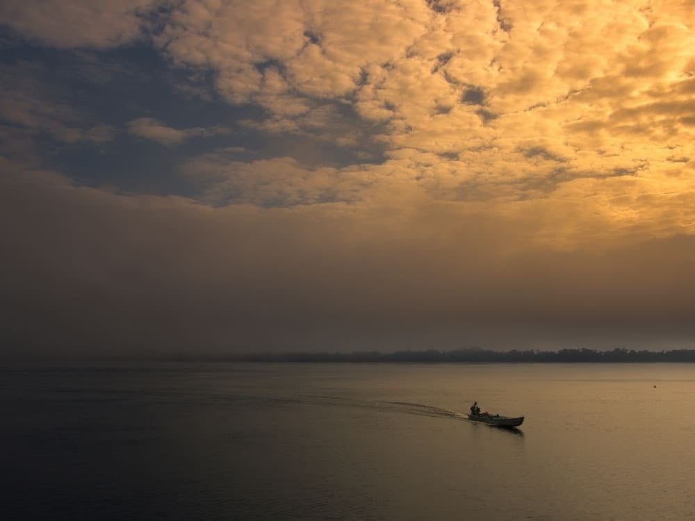 Una piccola canoa a motore che attraversa l'ampio Rio delle Amazzoni durante un'alba dorata, sotto un cielo nuvoloso. Una piccola canoa a motore che attraversa l'ampio Rio delle Amazzoni durante un'alba dorata, sotto un cielo nuvoloso.
